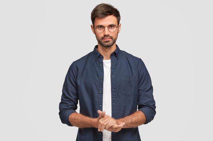 waist-up-portrait-handsome-serious-unshaven-male-keeps-hands-together-dressed-dark-blue-shirt-has-talk-with-interlocutor-stands-against-white-wall-self-confident-man-freelancer_273609-16320